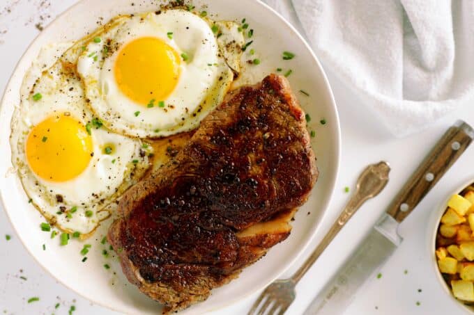 An overhead view of a plate of Steak and Eggs with a knife, fork, and bowl of hash browns near by. 