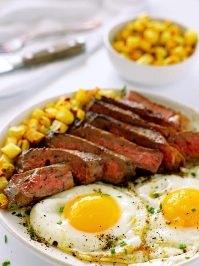 A plate of Steak and Eggs with hash browns on the side and a bowl of hash browns in the background. The steak is sliced into strips, showing that it is pink on the inside.
