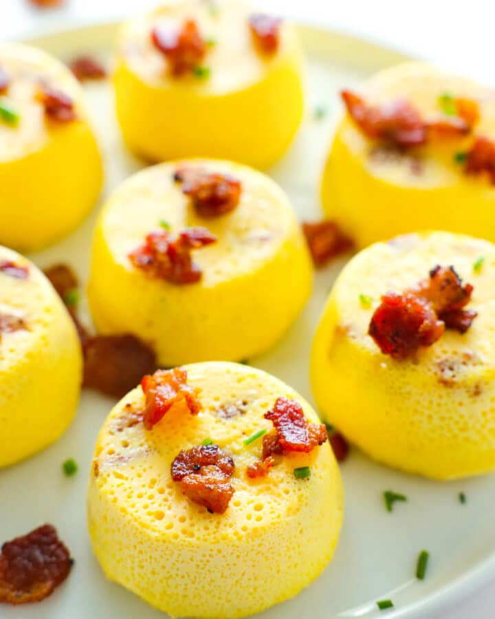 A close up of Egg Bites sitting on a white platter, pieces of cooked bacon and chives sprinkled on top.