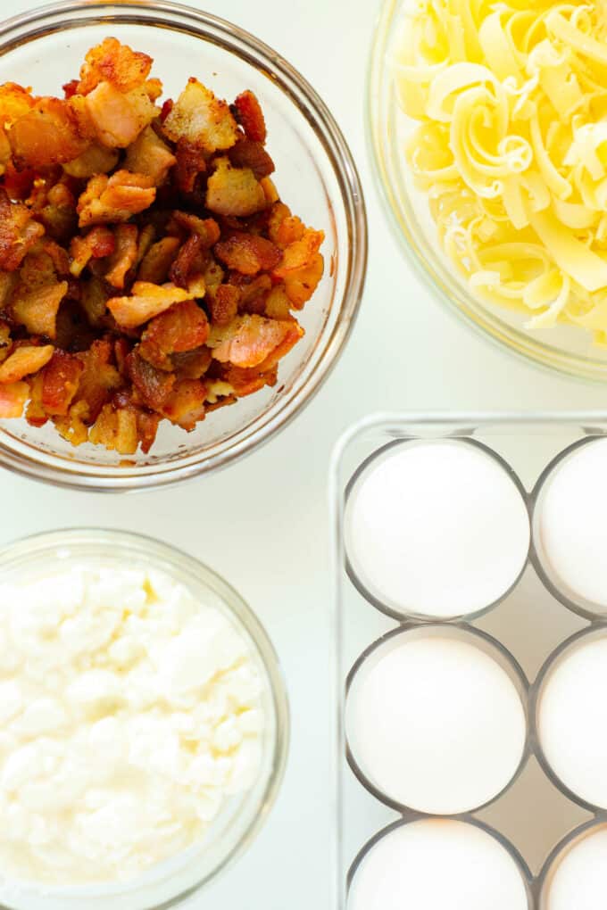 An overhead shot of eggs, cooked bacon, and freshly grated Monterey Jack and cottage cheese in separate bowls.