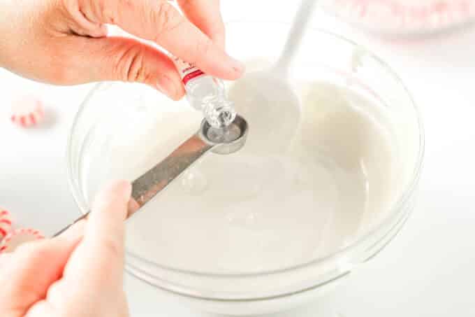 Peppermint oil being poured into a measuring spoon over the bowl of melted white chocolate wafers.