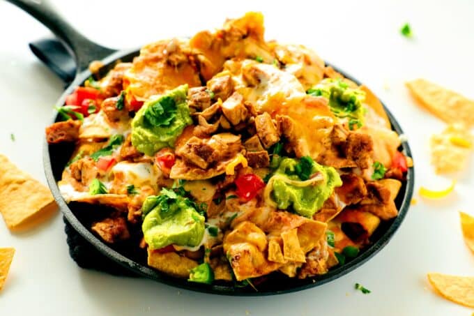 A pan of Chicken Nachos sits on the counter with chips scattered around it. The dish is topped with guacamole and diced tomatoes.