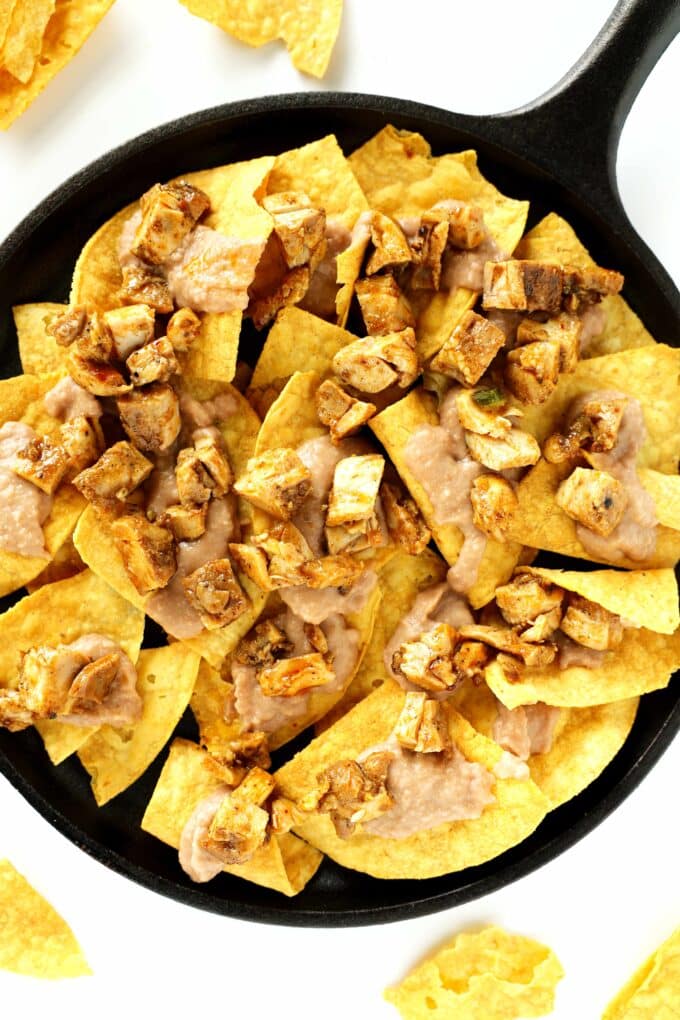 An overhead view of an iron pan with a layer of corn chips in the bottom and refried beans and diced chicken over top of them.
