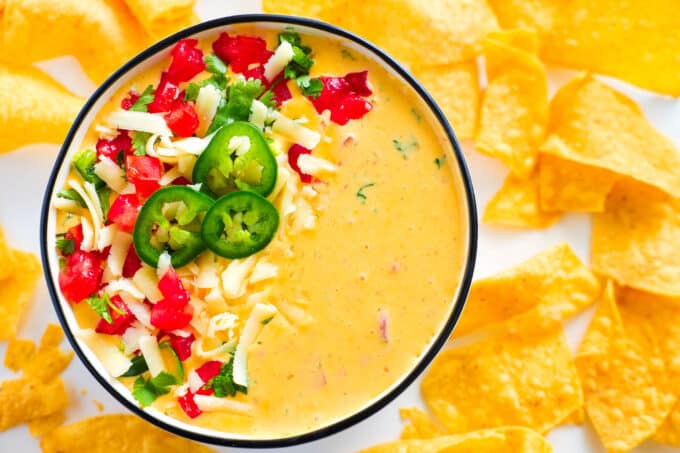 An overhead look at a bowl of Chile con Queso with fresh tomatoes, jalapeños, and cilantro sprinkled over one side of it and tortilla chips scattered all around.