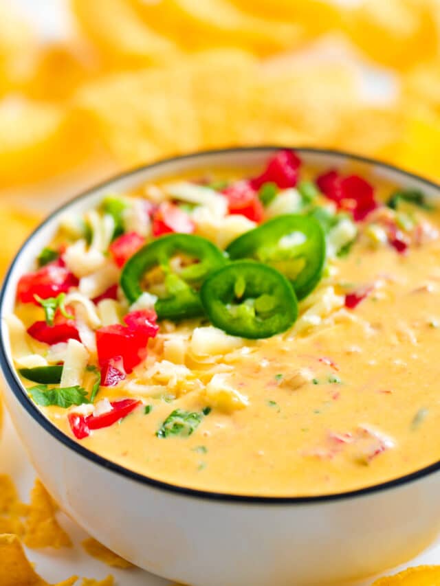 A close up of a bowl of Chile con Queso with a garnish of fresh tomatoes, jalapeños, and cilantro and tortilla chips sitting all around it.