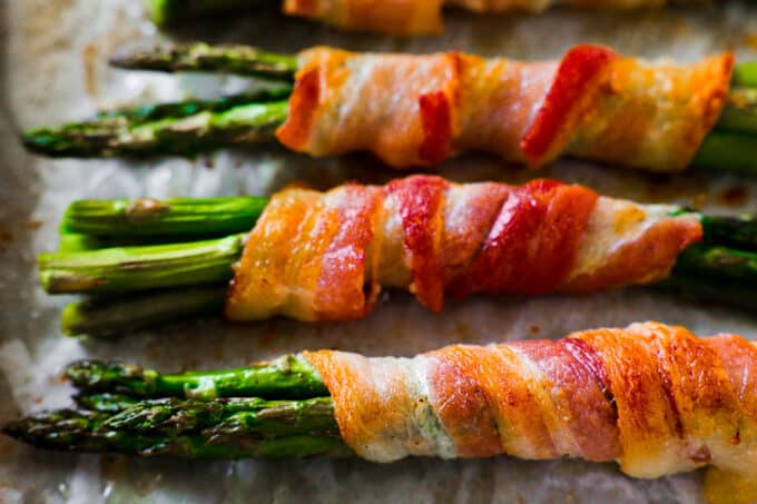 A close up, side view of Asparagus Wrapped in Bacon on a sheet pan. The asparagus is bright green and a little charred and the bacon is perfectly crispy.