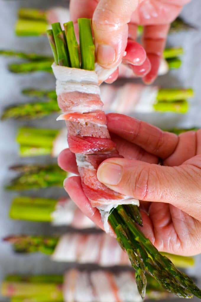 Hands wrapping a bundle of asparagus with a piece of raw bacon.