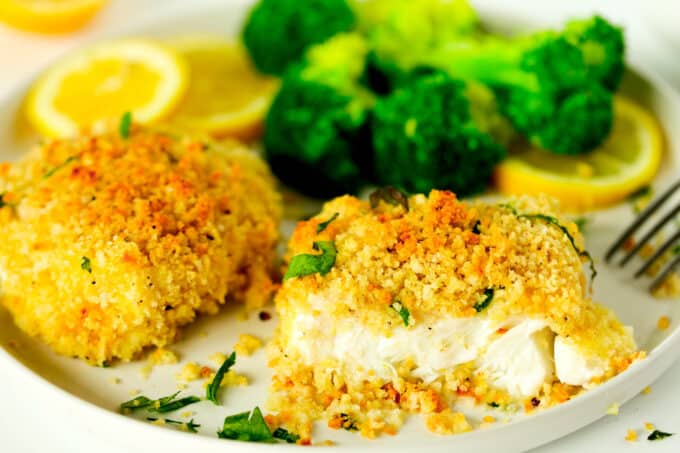 Two Baked Fish fillets on a plate with steamed broccoli and lemon wheels. One of the fillets is partially gone and the white, flaky fish is visible.