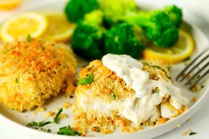 A plate with two fish fillets on it with tartar sauce on top of one of them. There is broccoli and lemon wheels on the back of the plate.
