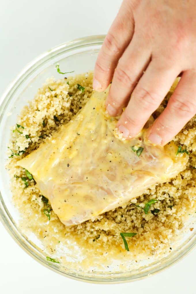 A hand pressing a fish fillet into the breadcrumb mixture.