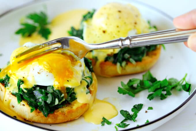 The side of a fork being used to cut into the runny yolk of one of the poached eggs in the Egg Florentine.