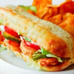 A Shrimp Po Boy on a plate with chips and pickles. You can see blackened shrimp, shredded lettuce, pickles, and tomato slices in the open side of the bun.