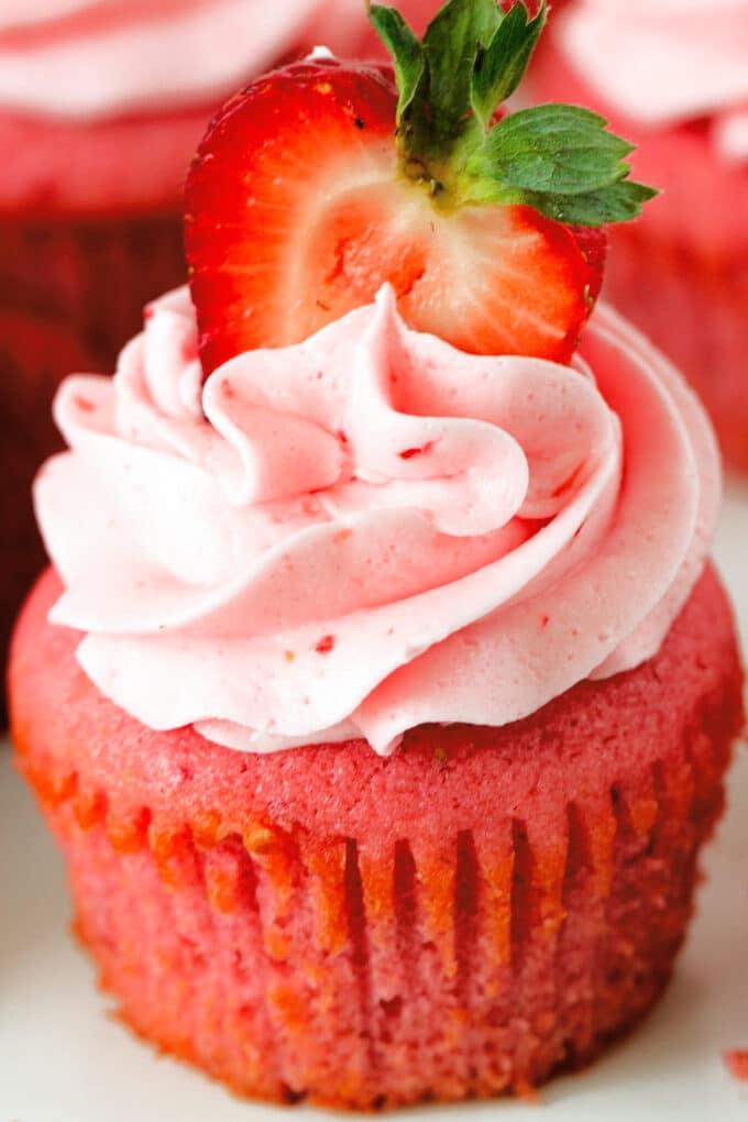 A close up of a Strawberry Cupcake with strawberry frosting and half of a fresh strawberry stuck on the top.