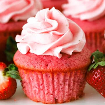 A close up of a Strawberry Cupcake with a swirl of strawberry frosting on top and fresh strawberries sitting next to it.