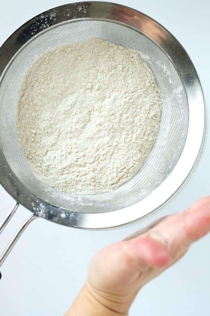 A hand patting the side of a fine strainer to shake the dry ingredients through.