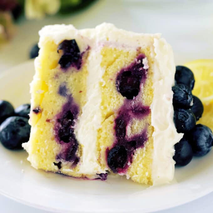 A piece of Lemon Blueberry Cake laying on a plate. The cake is yellow with large, purple blueberries throughout.