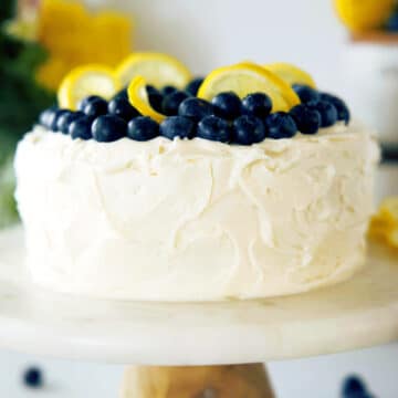 A side view of a Lemon Blueberry Cake on a marble cake stand. It is decorated with fresh lemon slices and blueberries.