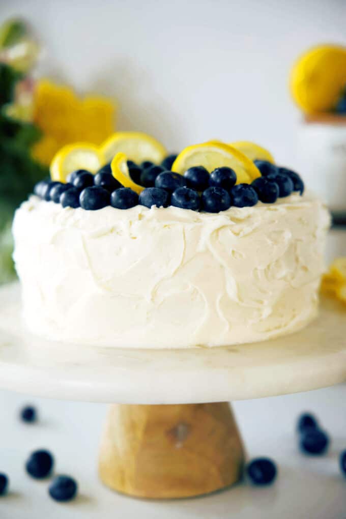 A side view of a Lemon Blueberry Cake on a marble cake stand. It is decorated with fresh lemon slices and blueberries.