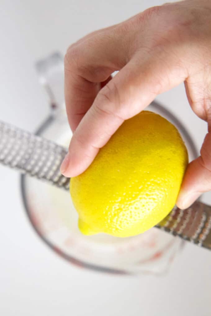 A hand zesting a lemon over a glass measuring pitcher.