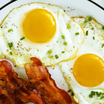 An overhead view of two sunny side up Fried eggs on a white plate with bacon on the side and chives sprinkled on top.