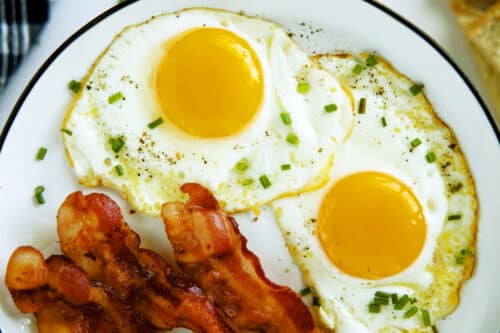 An overhead view of two sunny side up Fried eggs on a white plate with bacon on the side and chives sprinkled on top.