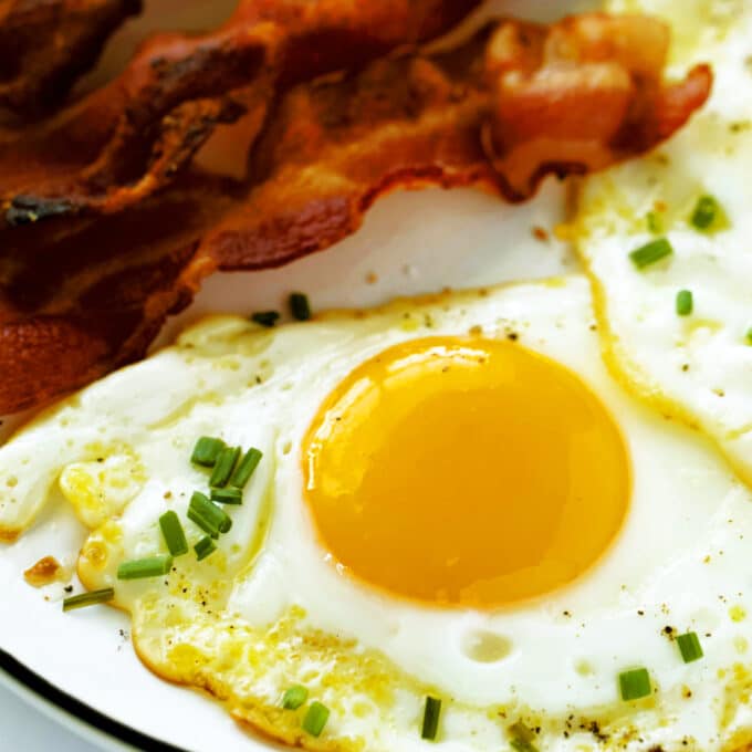 A close up of two Fried Eggs sprinkled with chopped chives. There are several pieces of bacon on the plate beside them.