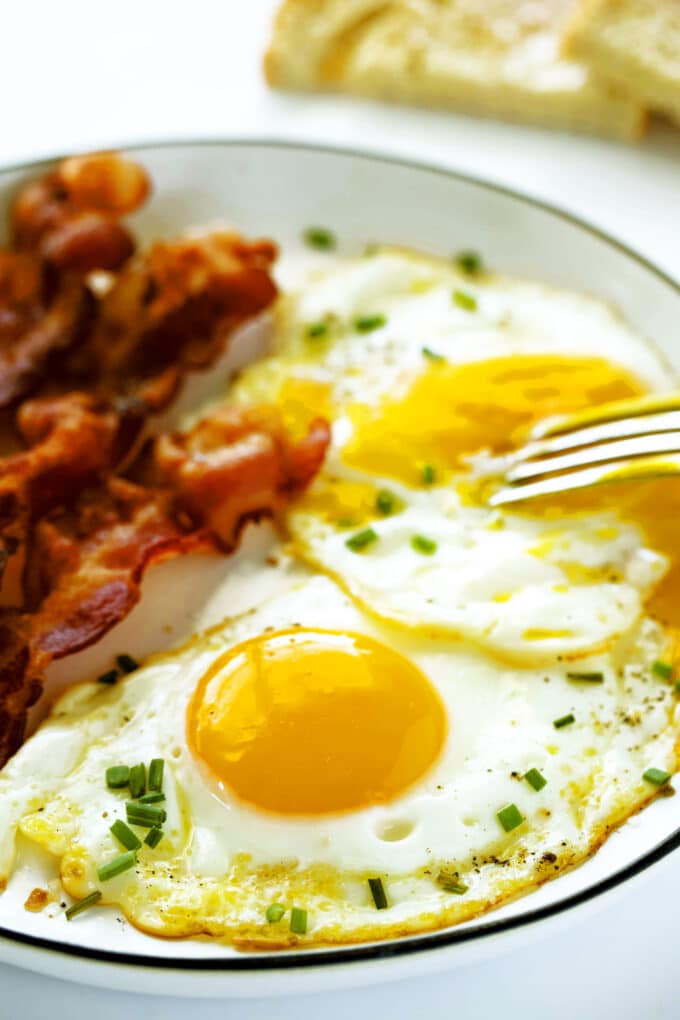 Two sunny side up Fried Eggs on a plate with bacon. A fork is being used to break one of the yolks.