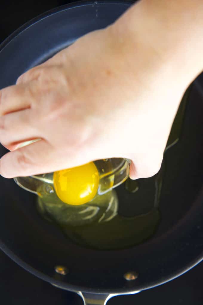 A hand gently sliding a raw egg from a small bow into the frying pan.