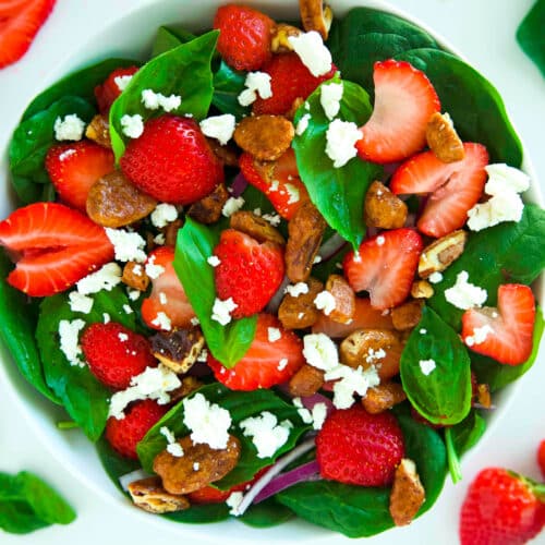An overhead look at a bowl of Strawberry Spinach Salad. The top is covered in sliced strawberries, candied pecans, and goat cheese crumbles.