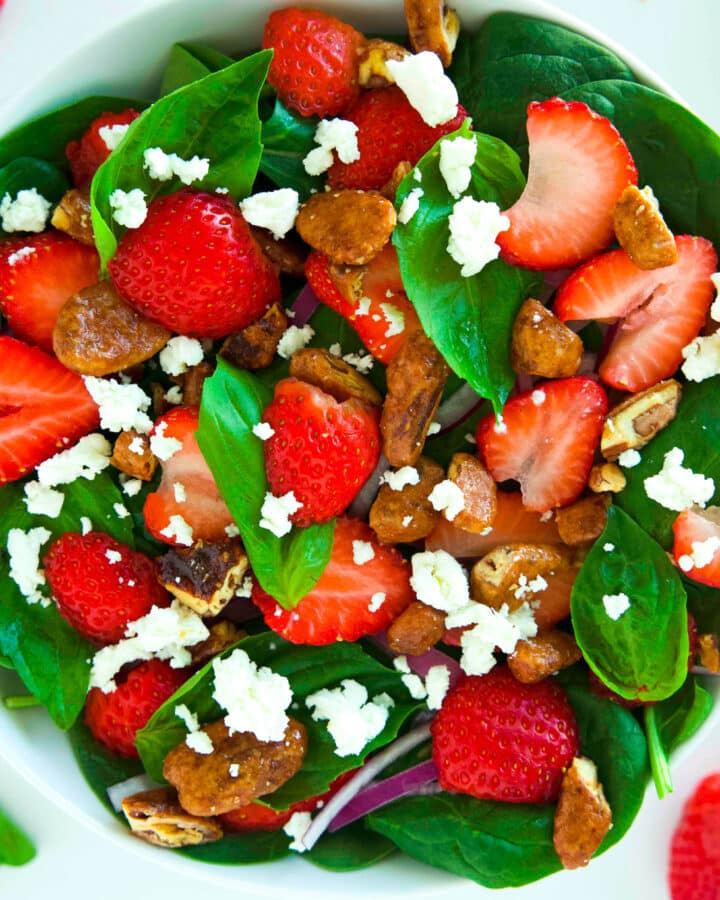An overhead look at a bowl of Strawberry Spinach Salad. The top is covered in sliced strawberries, candied pecans, and goat cheese crumbles.