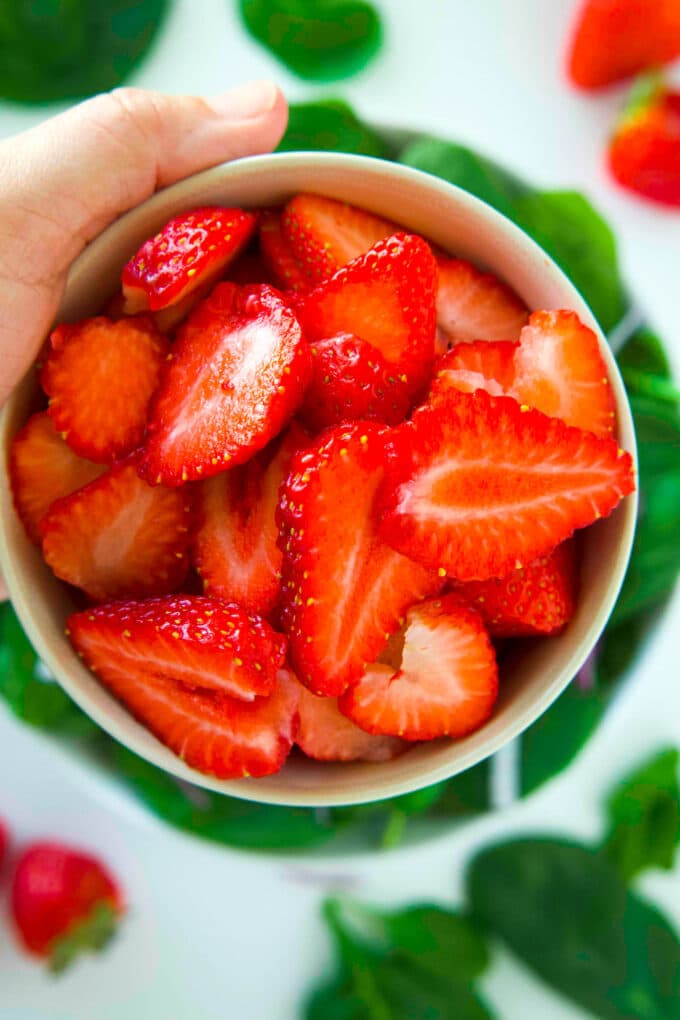A hand holding a bowl of sliced strawberries up to the camera. The are spinach leaves and whole strawberries sitting on the counter below.