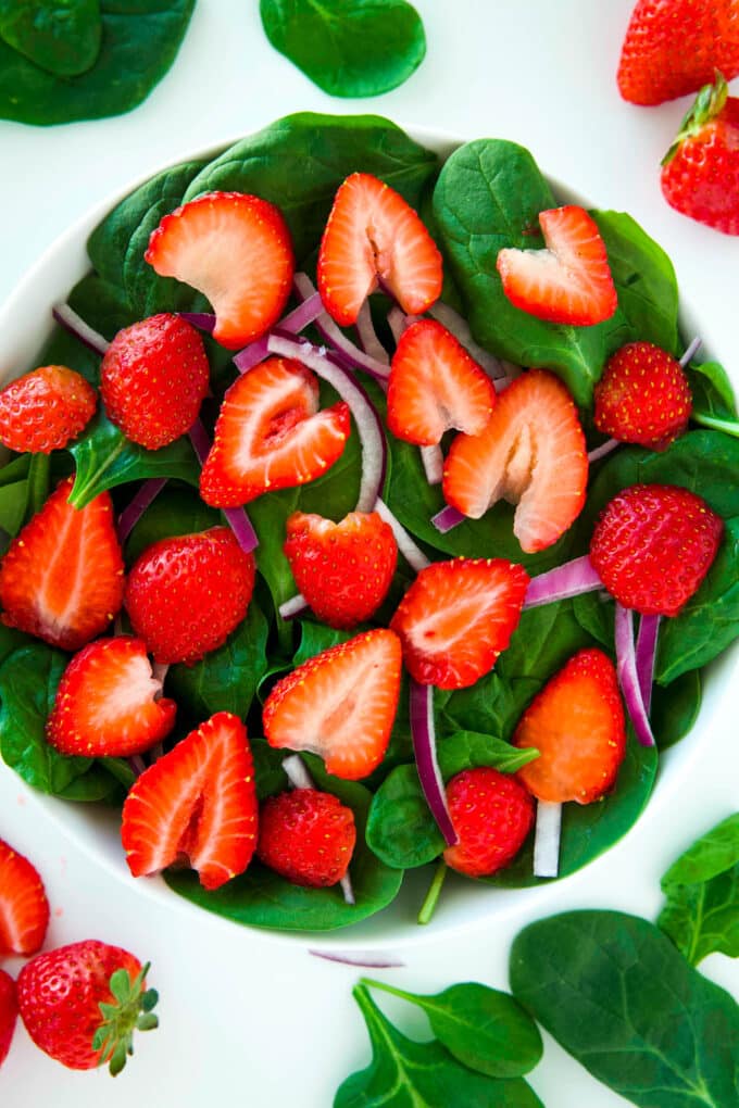An overhead view of a big bowl of baby spinach with sliced strawberries and red onion on top.