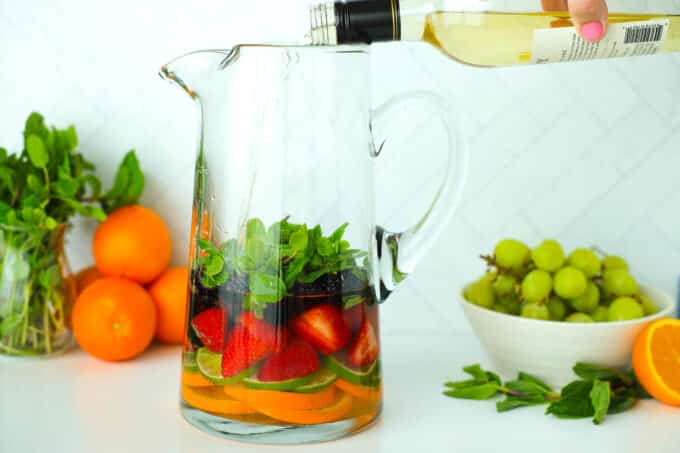 White wine being poured from the bottle into the pitcher of fruit and mint.