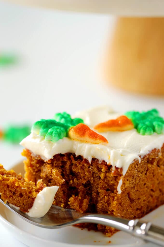 A piece of Carrot Cake with cream cheese frosting sits on a plate. There are carrot shaped candies on top and there is a fork laying next to it with a bite of cake on it.