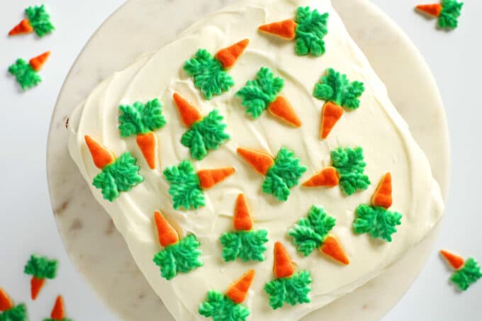 An overhead view of a whole Carrot Cake. It is square and iced with cream cheese frosting. There are little carrot candies all over the top and some sitting on the counter nearby.