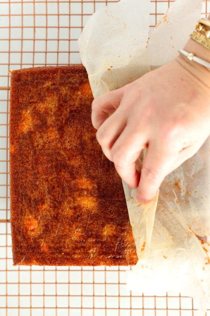 A hand pulling the parchment paper off the bottom of the cake that is sitting upside down on a cooling rack.