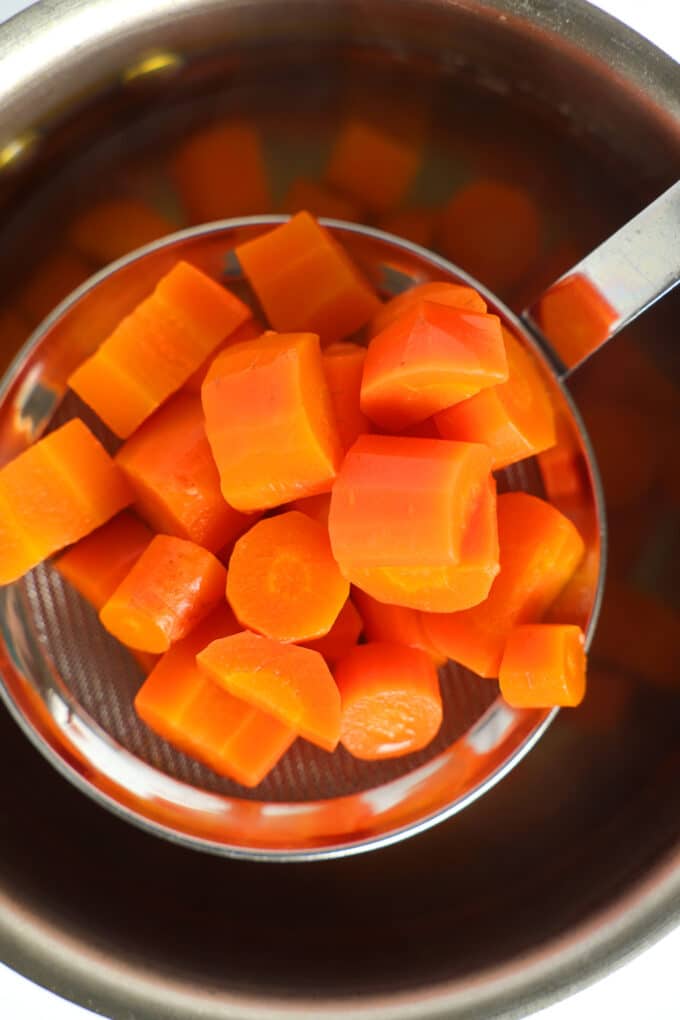 Chunks of cooked carrots being lifted out of the boiling water with a strainer.