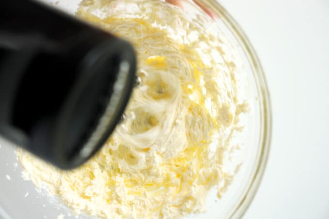 The cream cheese and butter being mixed with a hand mixer in a clear, glass bowl.
