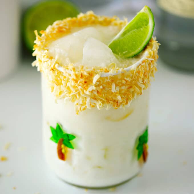 A close up of a Coconut Margarita. It is in a short glass with painted palm trees on it. The drink is white and there is a toasted coconut rim on the glass and a lime wedge garnish.