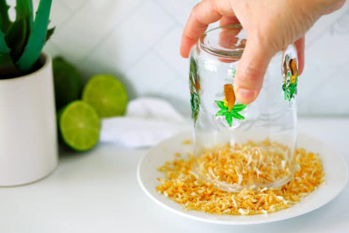 A hand twisting the rim of a glass in the toasted coconut on a small plate.