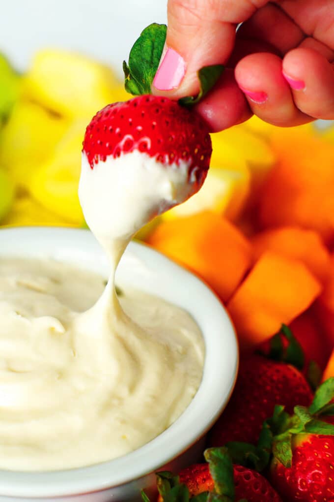 A hand dipping a strawberry into a bowl of Cream Cheese Fruit Dip that sits surrounded by an assortment of fruit.
