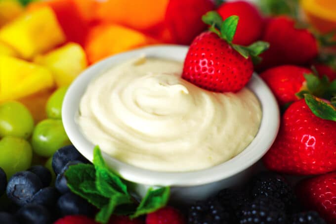 A bowl of Cream Cheese Fruit Dip sits in the middle of a platter of assorted fruits. A single strawberry is stuck in the dip.