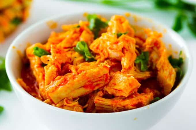 A bowl of Crockpot Buffalo Chicken sitting on the counter with fresh herbs laying in the background.