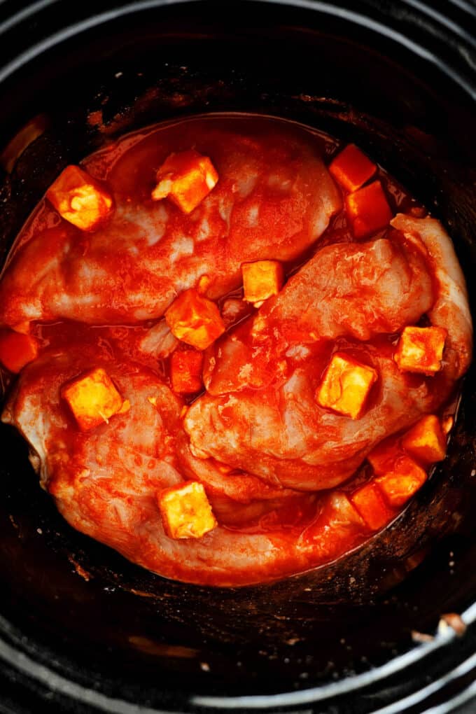 An overhead view of the raw chicken breasts in the crockpot. It's coated with hot sauce and seasoning, and has butter cubes sprinkled over it.