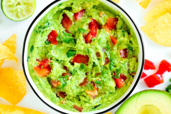 An overhead view of a bowl of Guacamole surrounded by tortilla chips, an avocado half, and diced tomatoes.