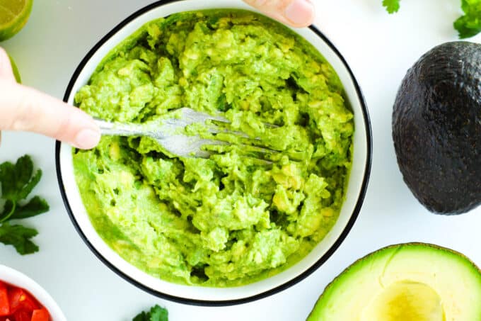 Avocado being mashed in a bowl with other ingredients laying nearby.