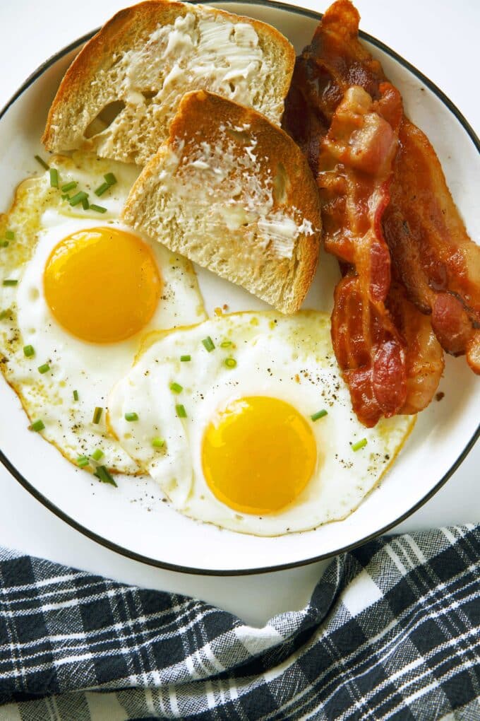 An overhead view of two Sunny Side Up Eggs on a plate with bacon and buttered toast. A plaid dish towel is laying next to it.