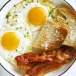 An overhead view of two Sunny Side Up Eggs on a plate with bacon and buttered toast.