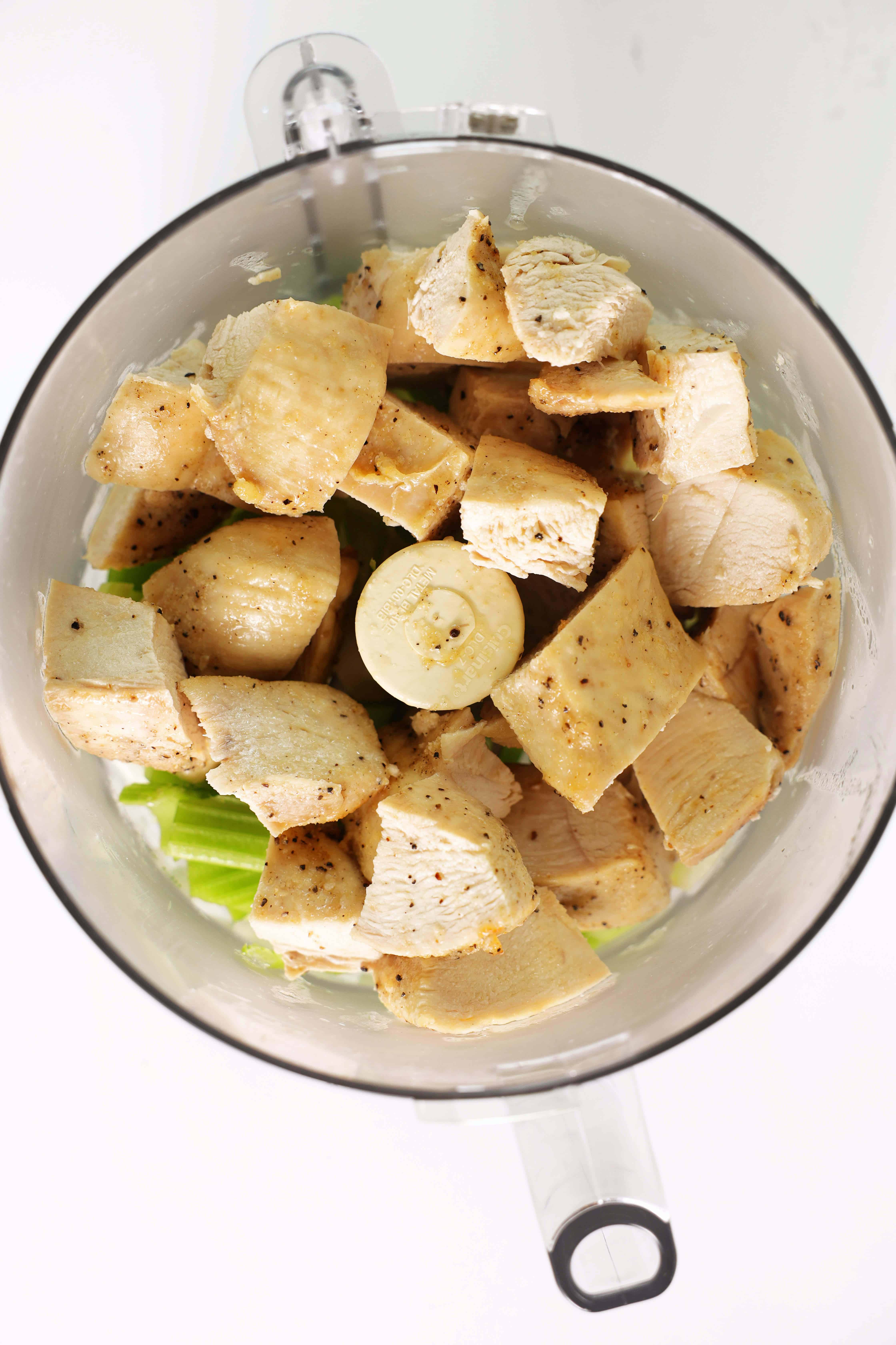 An overhead shot of cubed chicken breast and chopped onion and celery in a small food processor. 