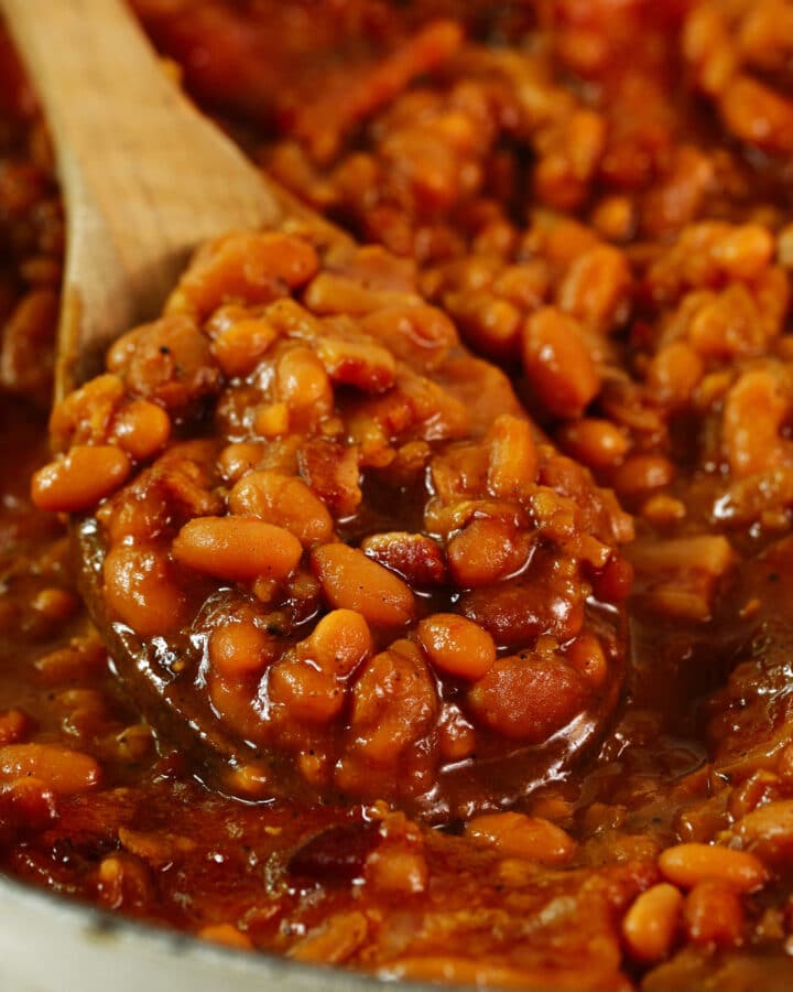 A close up of a wooden spoon scooping some Homemade Baked Beans out of the pot. The beans are in a thick, brown sauce.